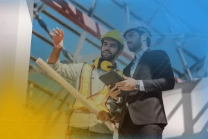 Um homem com vestes e capacete de construtor gesticula realizando explicações para outro homem de terno e capacete de construtor